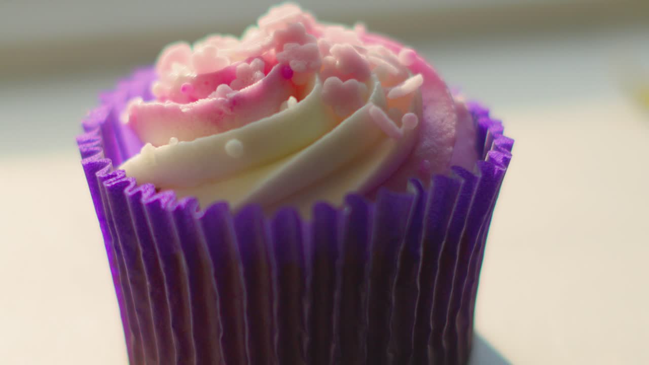 Pushing Shot Towards Cupcake in Natural Direct Sunlight with Deep Shadows on Window Sill with Purple and White Colored Frosting with Sprinkles.