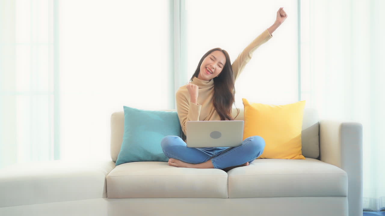 Relaxed Asian woman using her laptop computer sitting on the bed with her legs crossed, she is typing a message on the laptop keyboard and suddenly wins the prize, lottery, exited face expression