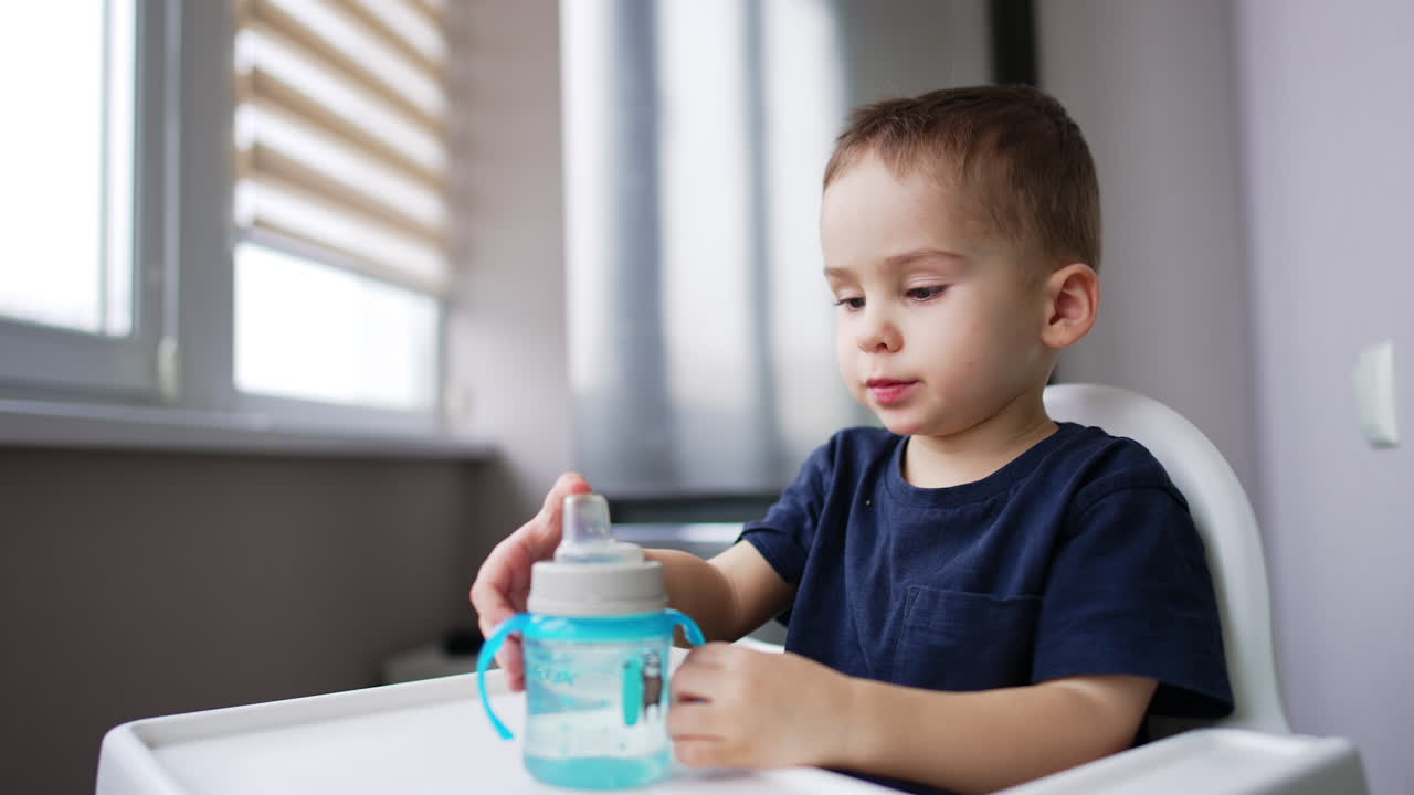 Adorable toddler drinking water from bottle. Baby boy sitting at high chair at home drinks.