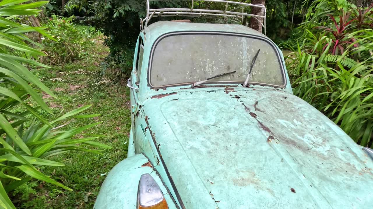 Old turquoise car sits abandoned in overgrown garden, natural daylight, slow forward camera movement