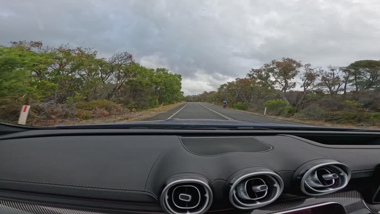 Interior view of a modern car driving a winding coastal road, with dashboard navigation screen visible. Overcast daylight, smooth camera movement, wide-angle perspective