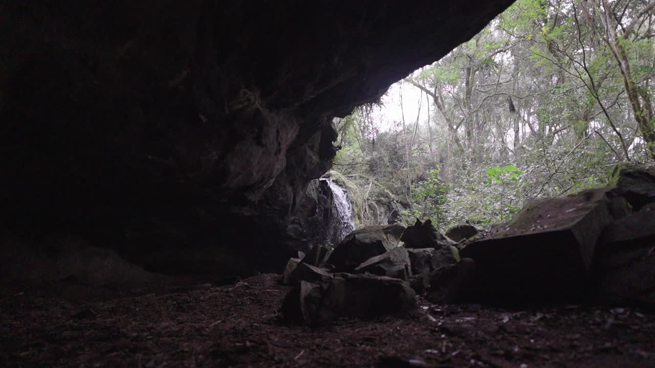 Dark cave grotto opens to a bright waterfall, with lush vegetation surrounding the rocky entrance, empty serene nature jungle backdrop