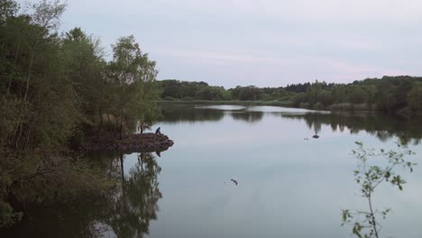 Weite-Panoramaaufnahme-Eines-Einsamen-Mannes,-Der-In-Der-Abenddämmerung-Nach-Sonnenuntergang-In-Der-Natur-Mit-Dem-Kopf-In-Den-Händen-Am-Rande-Eines-Wunderschönen-Kleinen-Sees-Sitzt-Und-Einen-Traurigen-Oder-Deprimierten-Eindruck-Macht