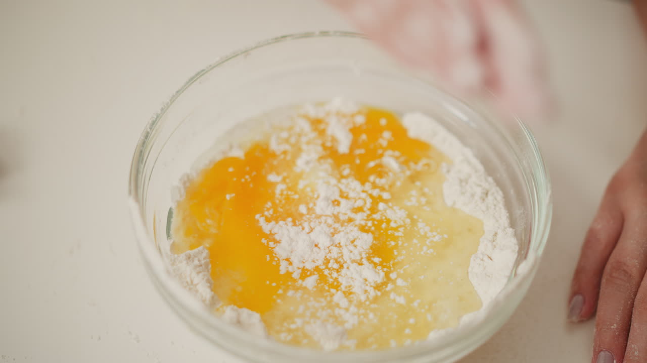 overhead view of flour with cracked eggs in glass bowl while powdered ingredient being dusted from above by hand, ready for mixing during cooking or baking process on clean kitchen surface