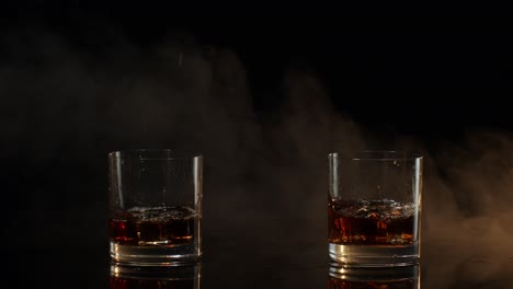 barmans dropping ice cubes into drinking glasses with whiskey, cognac, brandy on black background