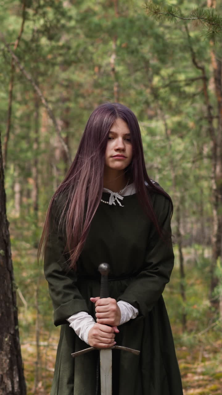 una adolescente seria con ropa tradicional de mediana edad clava una espada en el suelo en el bosque. una joven guerrera. reconstrucción de un evento histórico