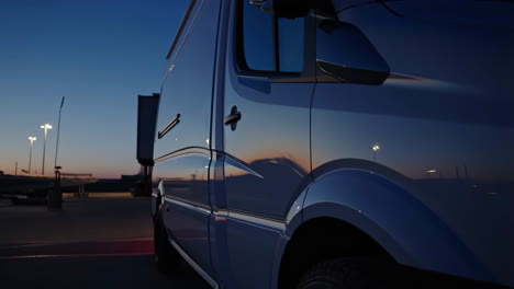 A blue van parked at dusk with streetlights in the background, showing reflections on its side