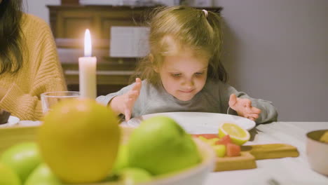 front view of a little girl picking up a piece of kiwi and eating it during a family dinner