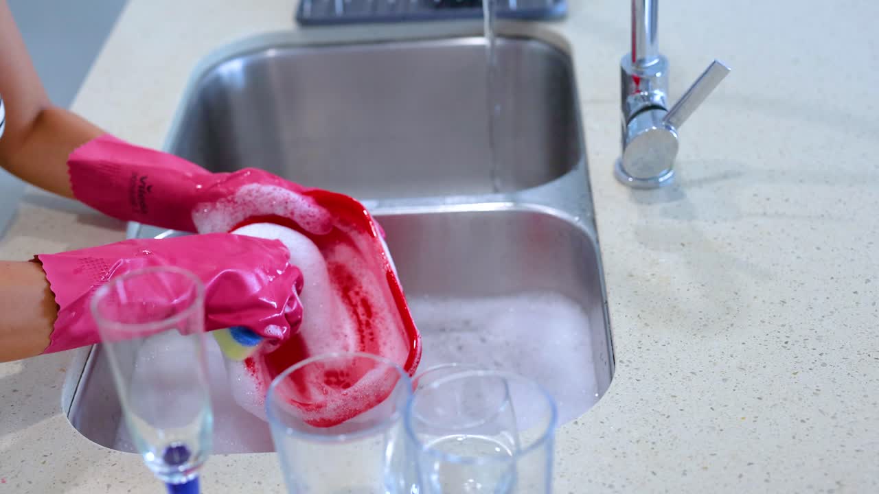 Hands in pink gloves washing a red dish in a bright kitchen with a stainless steel sink