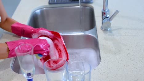 hands in pink gloves washing a red dish in a bright kitchen with a stainless steel sink