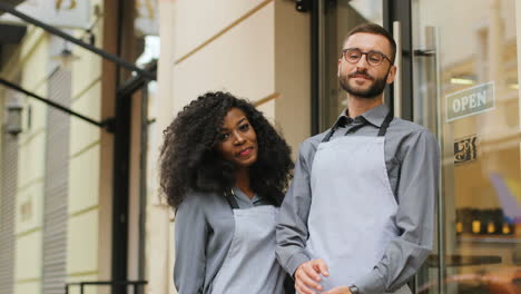 kaukasischer männlicher barista und afroamerikanischer weiblicher barista stehen an der tür eines modernen cafés, lächeln und schauen in die kamera