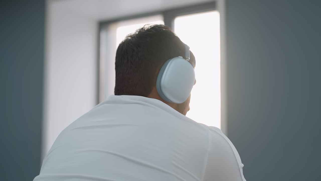 side view of young man wearing over ear headphone pedaling on stationary bike in bright gym space with sunlight streaming through window as he glances toward activity beyond exercise area