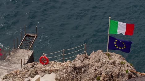 Italian and EU flags blowing on a concrete deck by rocky coast and ocean in Taormina, Sicily, Italy (Italia)