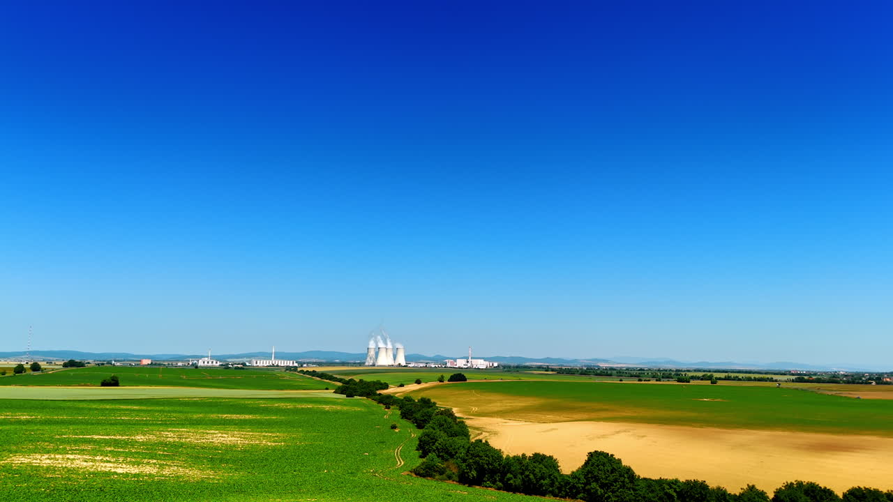 Flight over the vast plantation in the countryside. Wide tubes emitting white smoke are seen at backdrop. Aerial view.