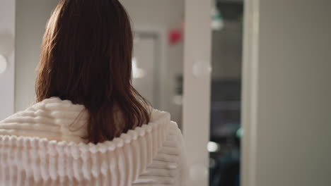 woman stands in spacious bathroom. brunette woman in bathrobe with hood after beauty treatments stands near mirror with lamps in bathroom backside view