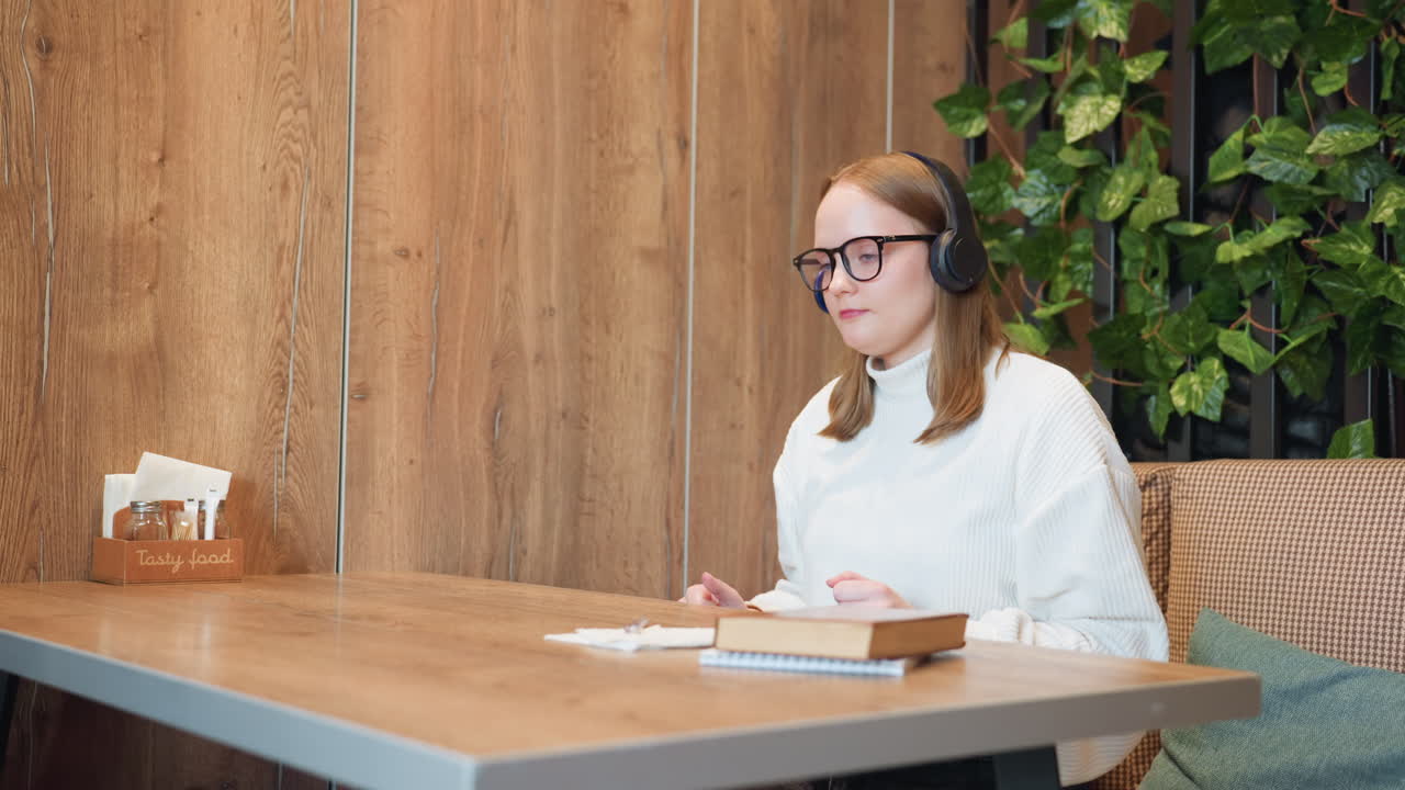 Woman in white sweater adjusts headset while sitting at wooden table with book and napkin sketches, preparing to enjoy music in cozy indoor space decorated with plants and soft furnishings