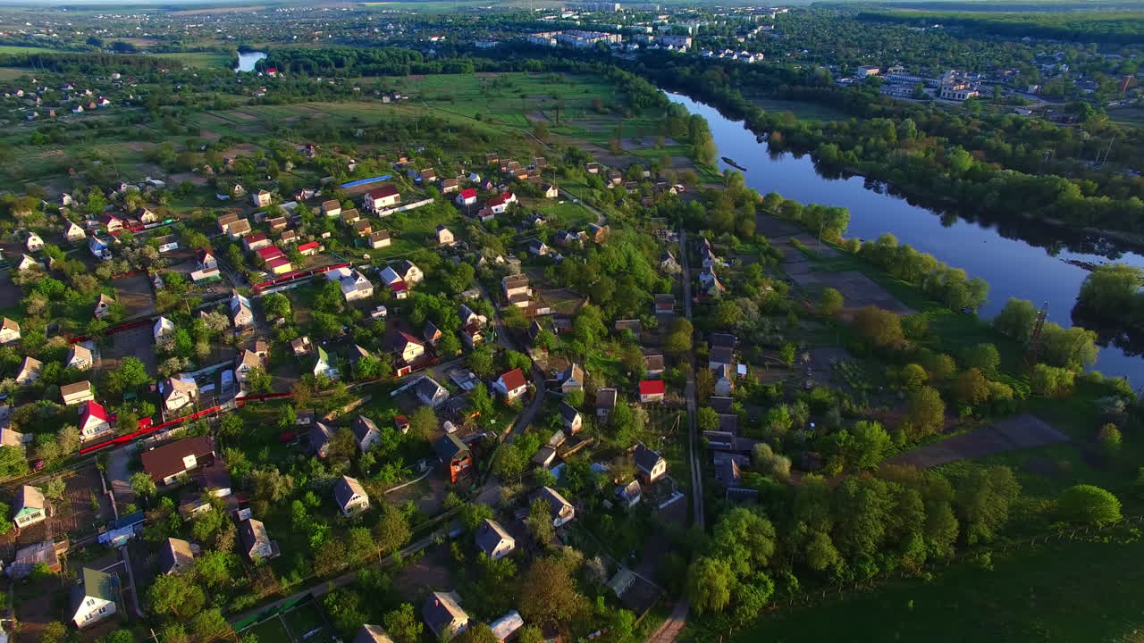Flight over the rural houses at the river bank. Vast green city panorama at backdrop of blue clear sky at dawn. Top view.