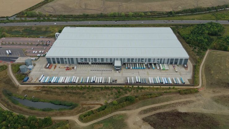Luftfoto i fugleperspektiv af et travlt distributionscenter i Bedford, England, med lastbiler og trailere, der læsser varer af ved store læsseramper
