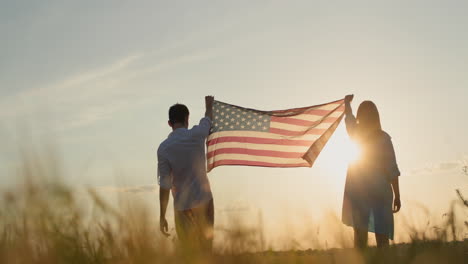 una pareja levanta la bandera de estados unidos sobre un campo de trigo mientras se pone el sol. concepto del día de la independencia de los estados unidos. video en 4k