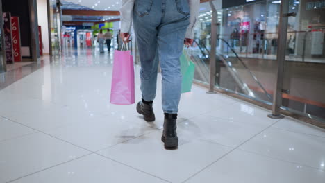 back view of a stylish woman walking confidently through a modern shopping mall, carrying pink and green shopping bags in both hands, she strides through the busy, well-lit retail space