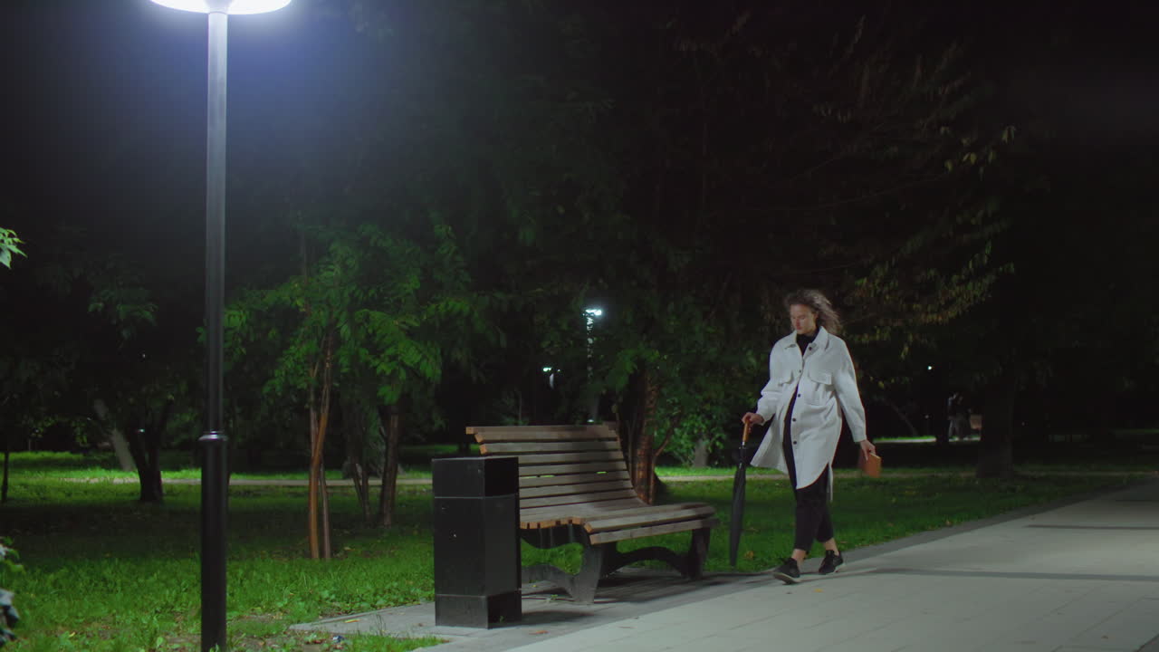 Student walks to wooden bench holding umbrella and book under glowing streetlight surrounded by trees with leaves swaying and falling in breeze prepares to sit and read quietly