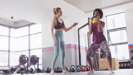 Video-of-diverse-female-fitness-trainer-instructing-woman-doing-squats-with-kettlebell-weight-at-gym