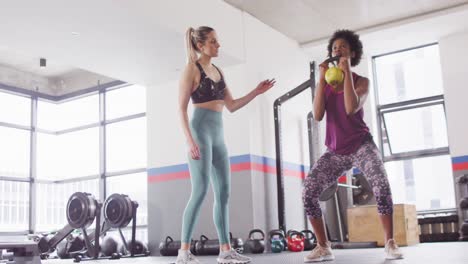 video de una entrenadora de fitness femenina diversa instruyendo a una mujer haciendo sentadillas con peso de kettlebell en el gimnasio