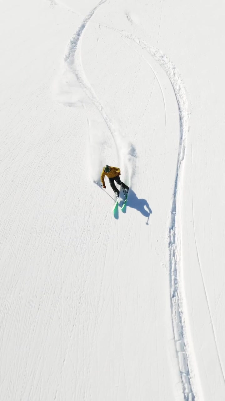 An exhilarating FPV drone shot capturing a skier carving through deep powder snow on a bright winter day. The dynamic perspective highlights the skier's movement and the untouched snowy terrain