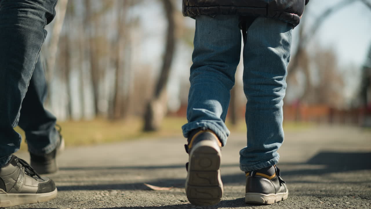 A leg view of an adult and a child walking on a pavement, the adult is wearing jeans and boots, while the child is dressed in a black leather jacket, jeans, and boots, with a blurred background