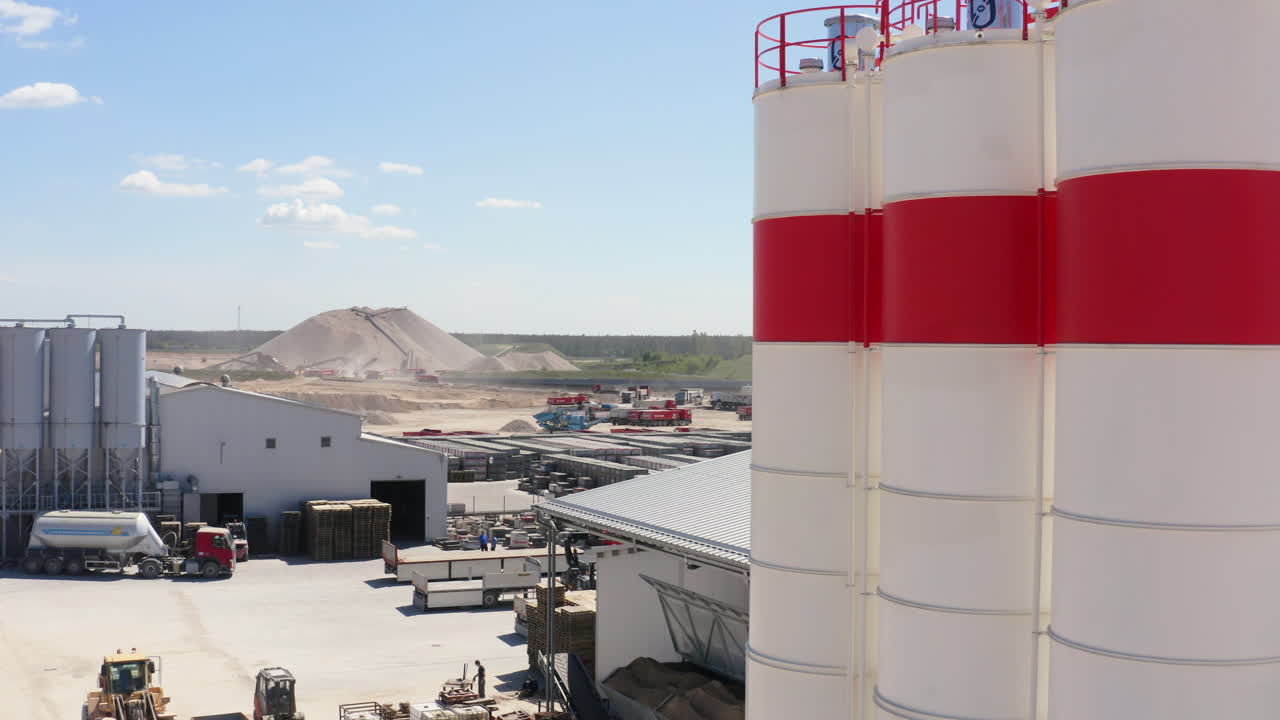 Industrial Complex With Large Silos, Storage Buildings, Trucks, And Piles Of Raw Materials On Sunny Day. ascending drone shot