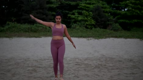 indian woman in pink sportswear exercises at the beach, standing leg stretches
