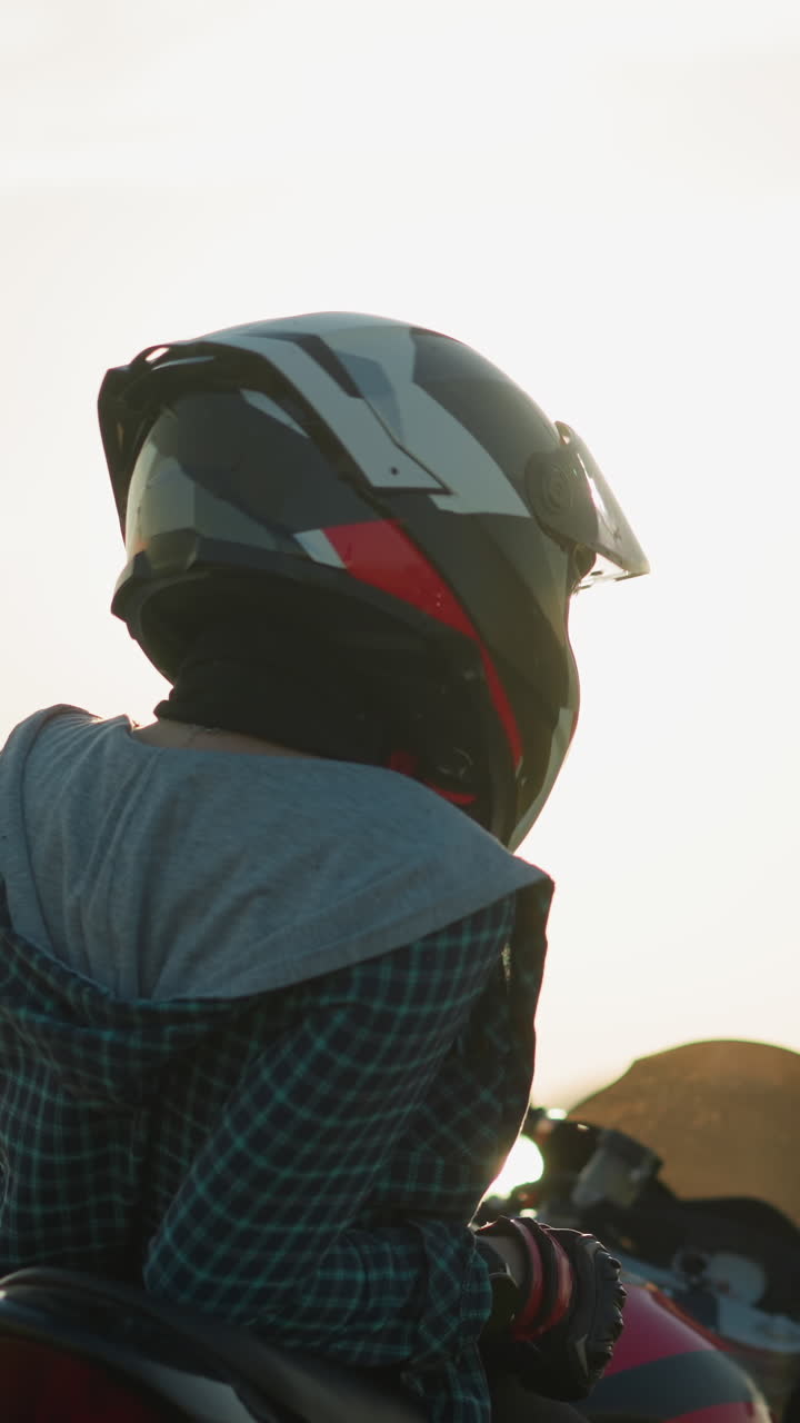 Back view of a lady relaxing on a red power bike in an open field, her helmet visor open as she gazes into the distance, with a scenic view of water and fields in the background