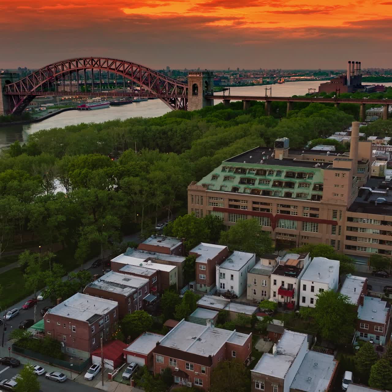 Lovely green residential area near the river waterfront. Hell Gate Bridge at the backdrop against cityscape and orange sky