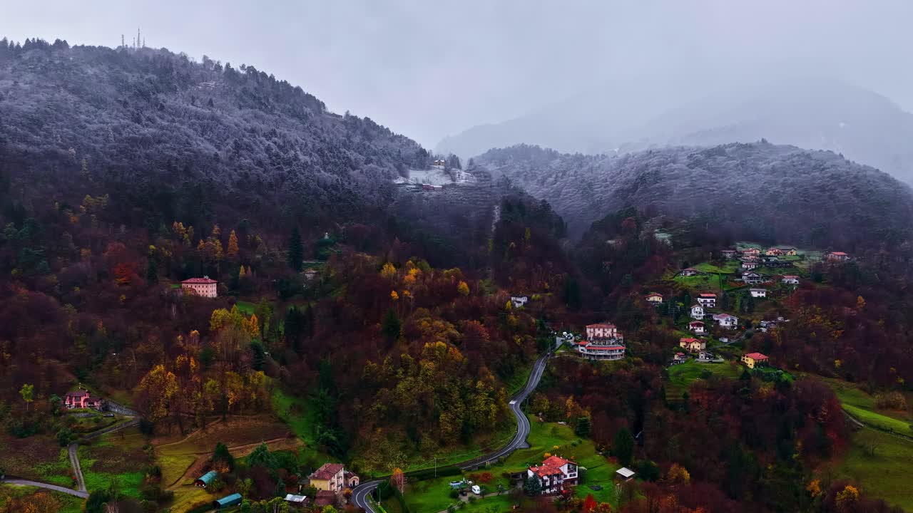 Nevicata su un piccolo villaggio rurale italiano circondato da colline naturali innevate e una foresta dai colori autunnali, vista aerea