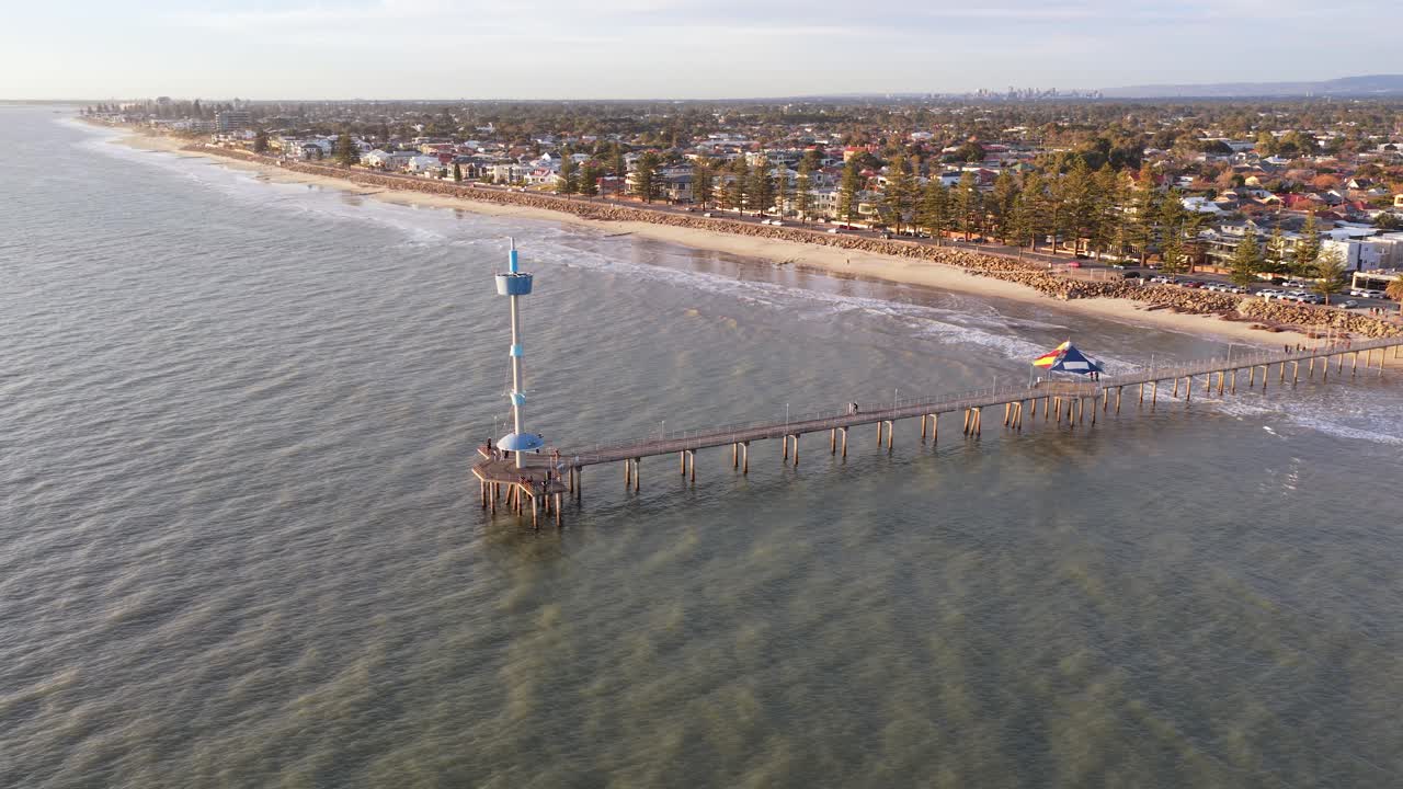 Wide drone orbit of Brighton Beach jetty and waves, revealing Adelaide's suburbs and city, South Australia