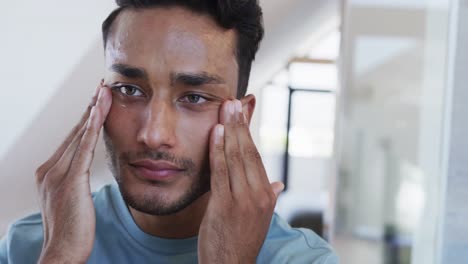 Smiling-biracial-man-looking-in-mirror-and-applying-face-cream-in-bathroom,-slow-motion