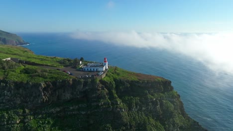 Drone-Empuja-Hacia-El-Faro-Mirando-Al-Océano-Azul-Con-Nubes-Brumosas-Iluminadas-Por-El-Sol-En-El-Horizonte