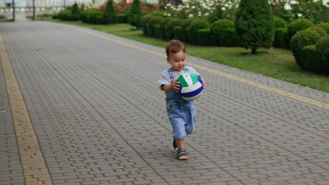 caucasian toddler in jeans romper playing with a ball in the park. happy child picks up a ball and runs with it approaching camera.
