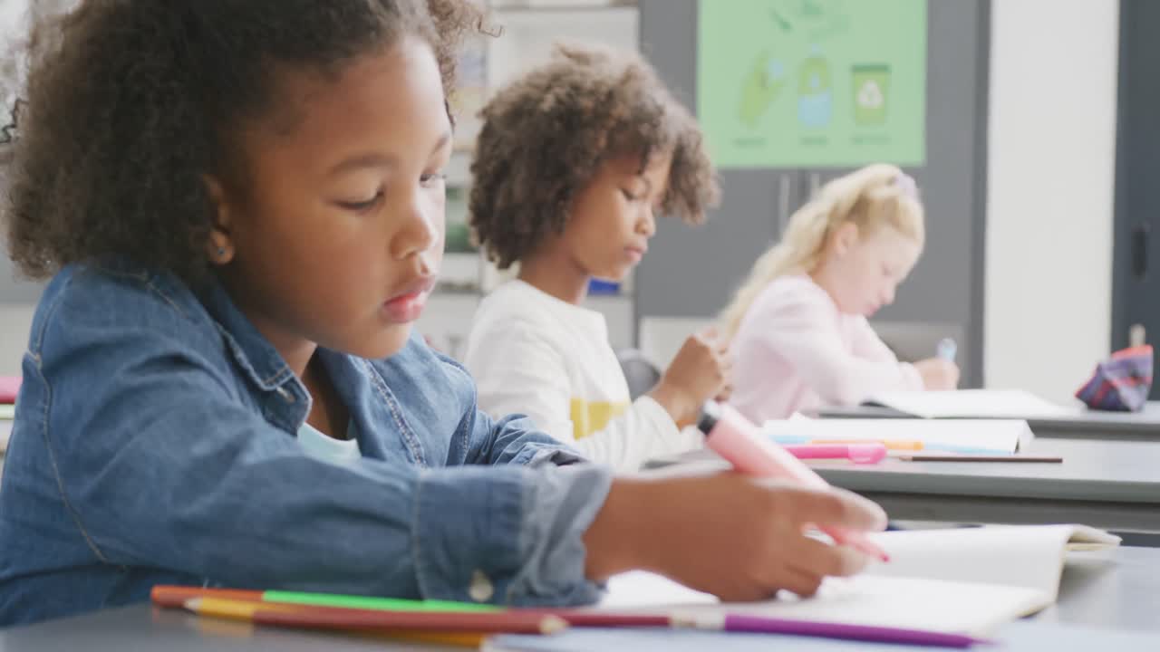 Video of biracial schoolgirl sitting at desk writing in diverse school class