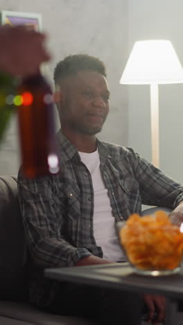 young black man carries bottles of cool drinks to enjoy football championship with best friends in apartment. guy with laptop sits checking score