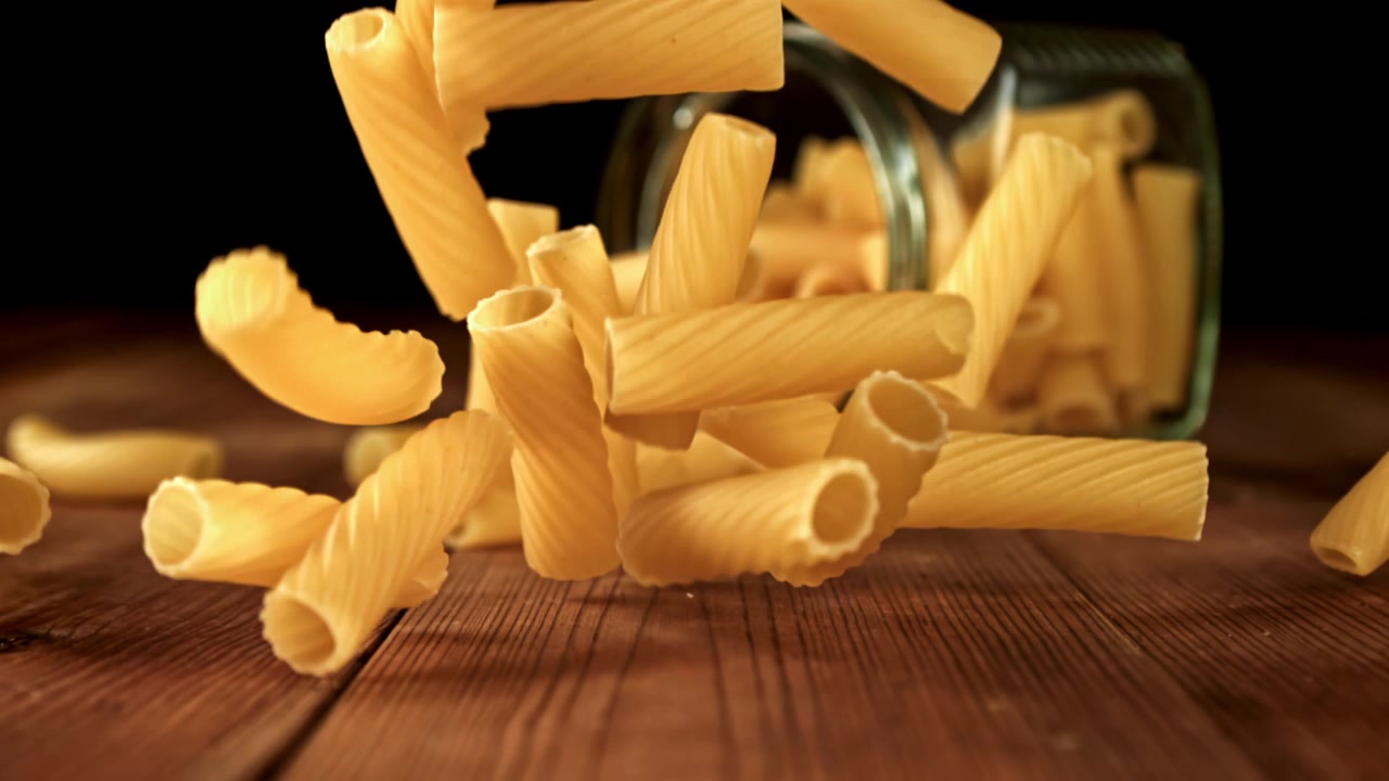 Pasta rigatoni secca che cade da un barattolo su un tavolo di legno al rallentatore
