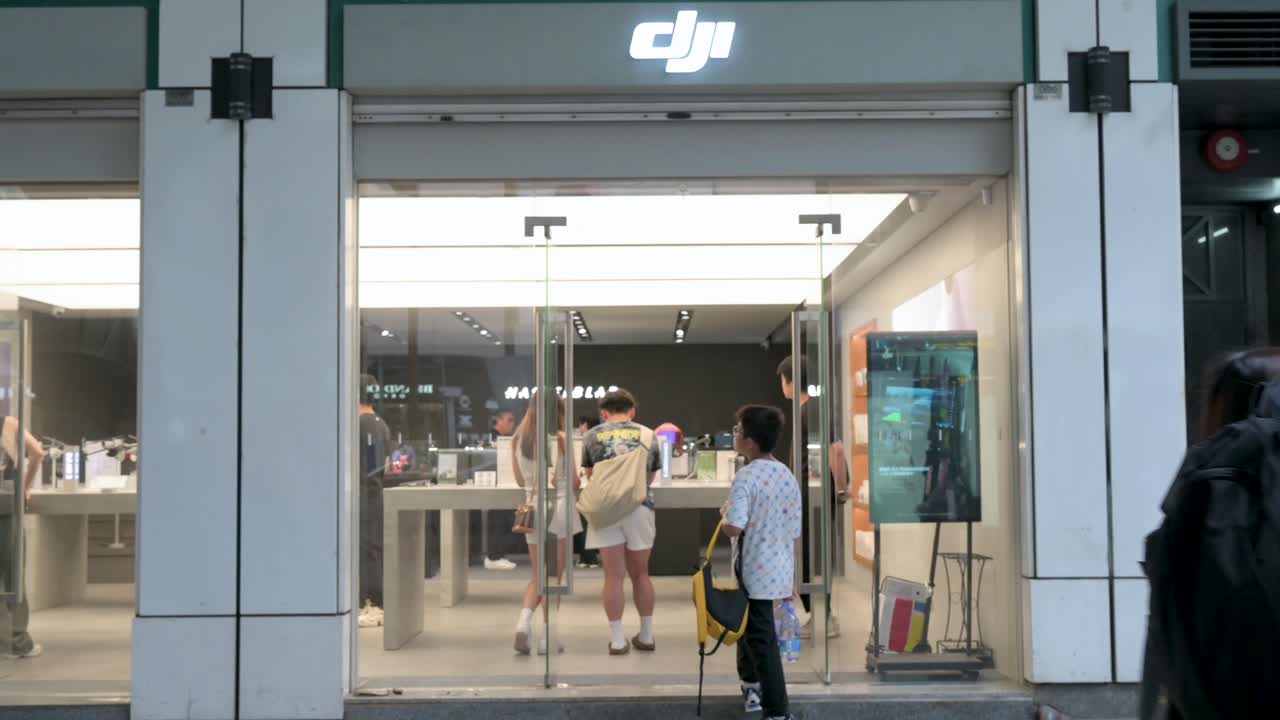 Customers browse DJI’s high-tech drone and camera gear offerings inside the store, while pedestrians pass by the illuminated Hong Kong storefront at night.