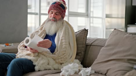 an elderly man with a thick gray beard in a red cap wearing a white plaid and blowing his nose in a napkin while he is sick and has a runny nose and sits at home on a modern sofa in the apartment