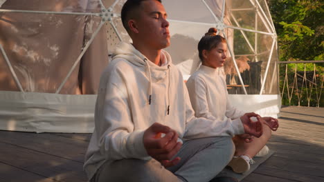 Man and woman sit in lotus poses at glamping. Concentrated couple of yoga practitioners meditates at eco resort retreat. Body and mind harmony
