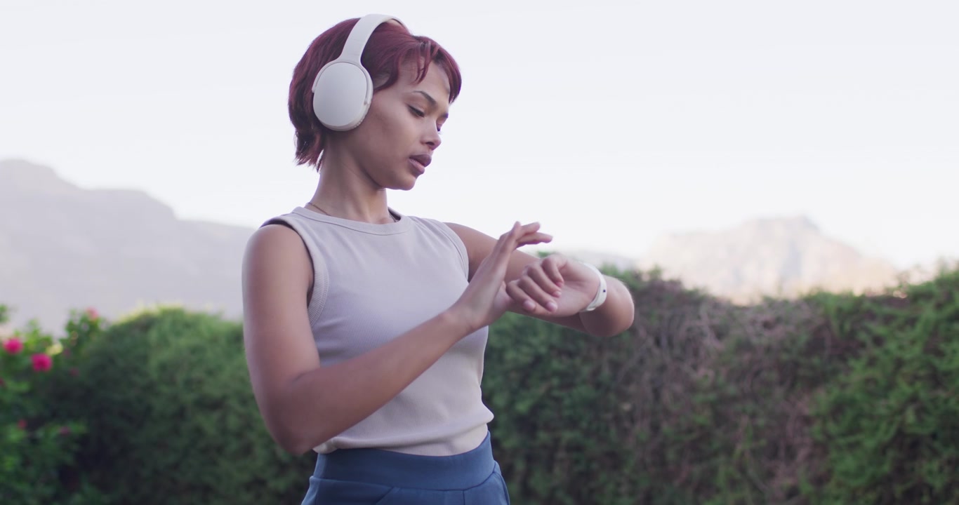 African American woman lifting headphones in yard adjusting tapping white watch preparing run