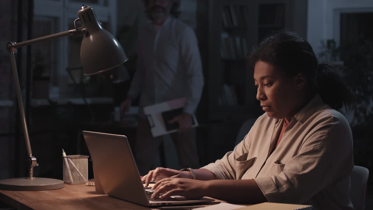 Woman working late on a laptop in an office setting, interacting with a colleague