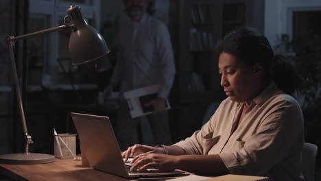 woman working late on a laptop in an office setting, interacting with a colleague