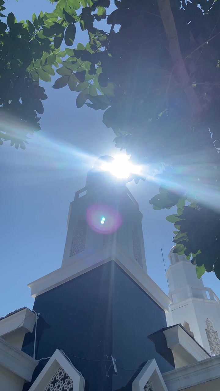 Pancaran Matahari Terang di Balik Menara Masjid Putih yang Dibingkai Daun Hijau