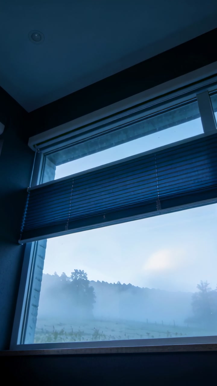 Vertical video: Showing window view with dawn revealing fog lifting over field in room, with blind