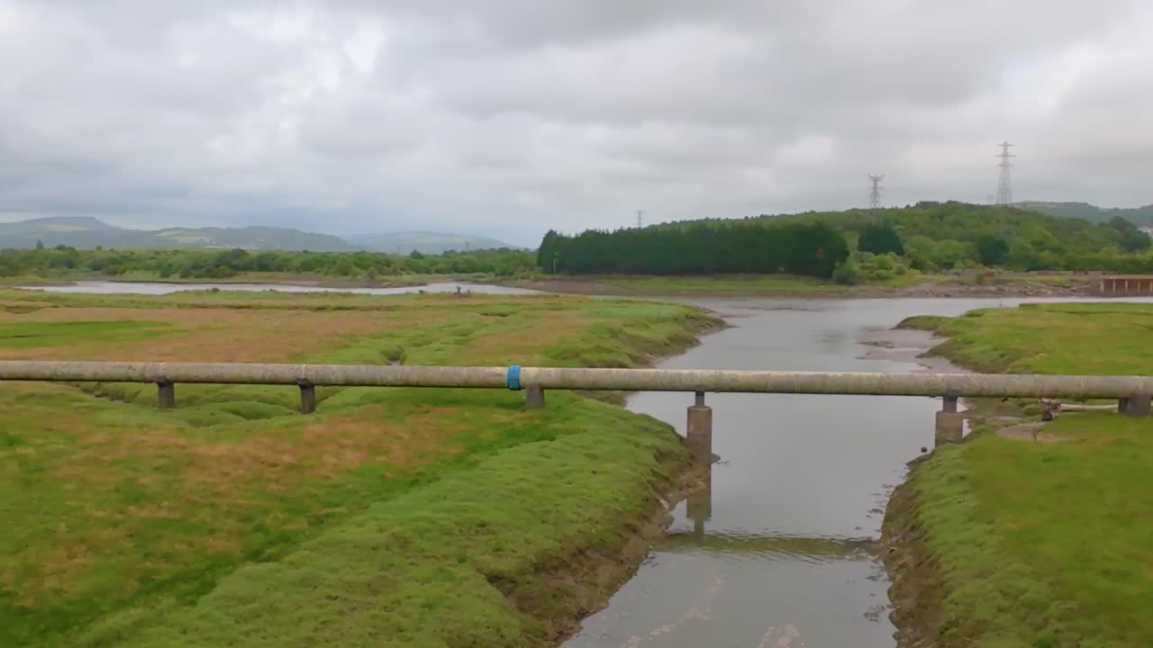 Gas Energy Pipeline on Marshland Near River with Forest Plantation and Electricity Pylons in Distance. Pipe near River Neath in South Wales.